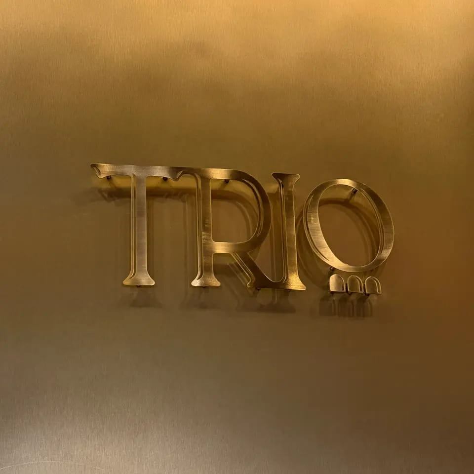 The TRIO logotype in raised brass letters on a warm brass wall
