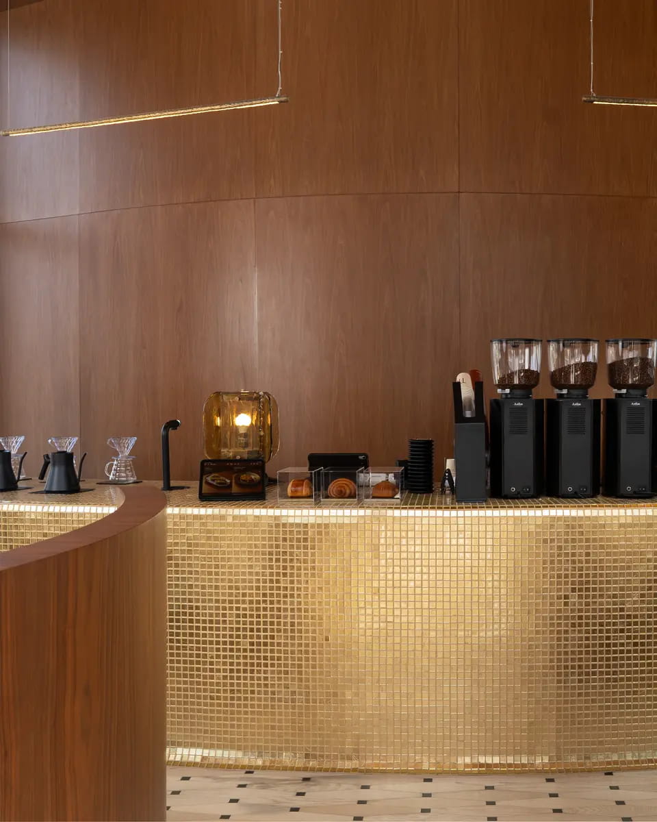 The espresso bar: a curved counter clad in gold mosaic under a walnut wall, pour-over kettles, a glowing amber lamp, pastries and three grinders along the top