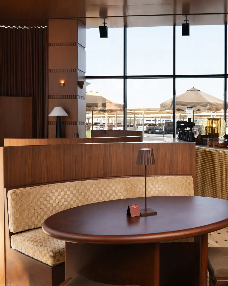 A curved booth in woven sand upholstery around an oval walnut table with a small table lamp, the terrace umbrellas beyond the tall windows