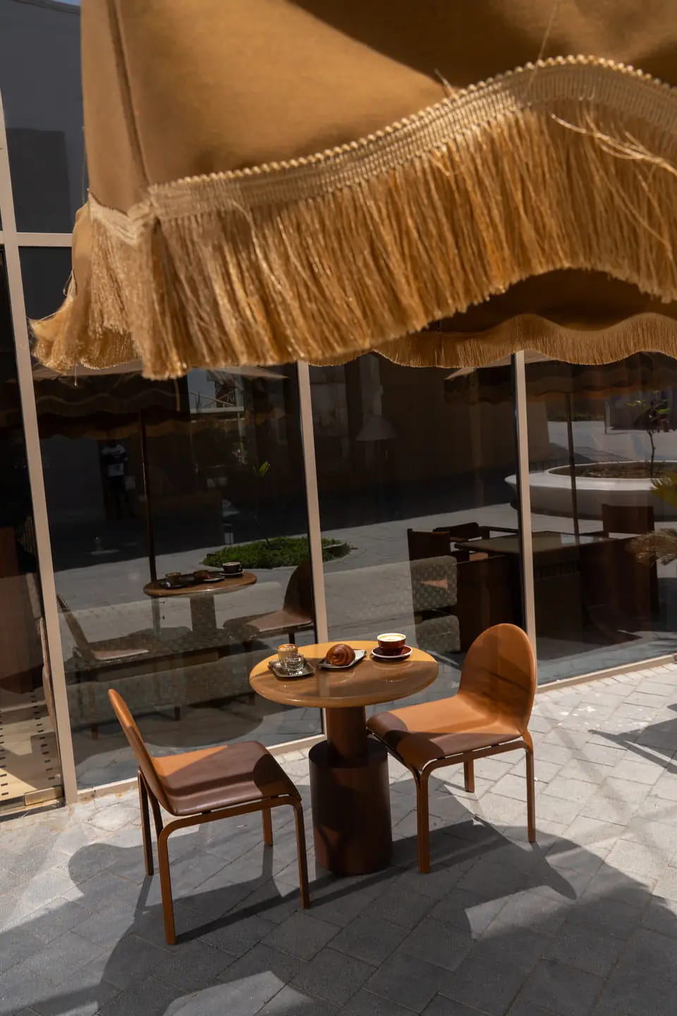 Coffee and a croissant on a round terrace table with two caramel leather chairs, the fringe of a sand umbrella overhead in bright sun