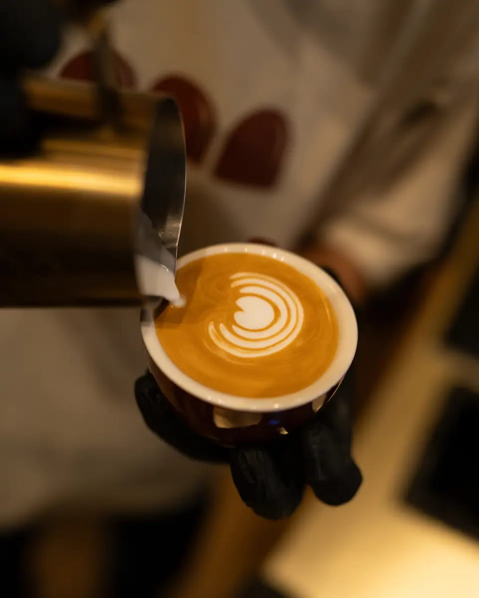 A gloved hand holds a latte with tulip art beside the steaming jug