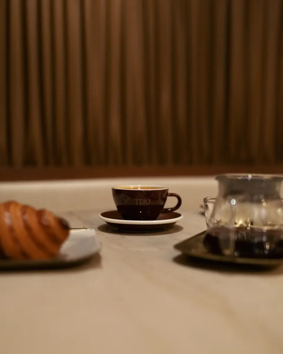 A cup of espresso, a pastry and a glass teapot on a white marble table against pleated drapery
