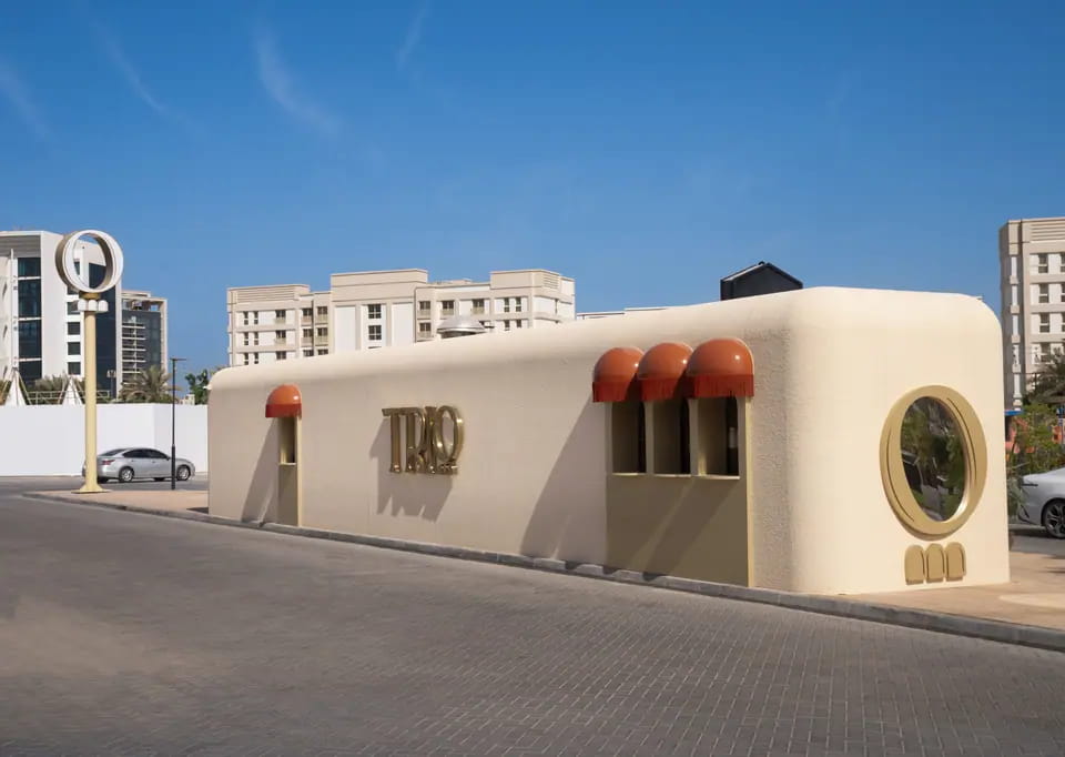 TRIO's soft-cornered sand-coloured container at Lagoon Stop under a clear blue sky: a brass TRIO wordmark, three fringed terracotta awnings over arched windows and a round brass-framed porthole, with the O-and-arches pylon beyond