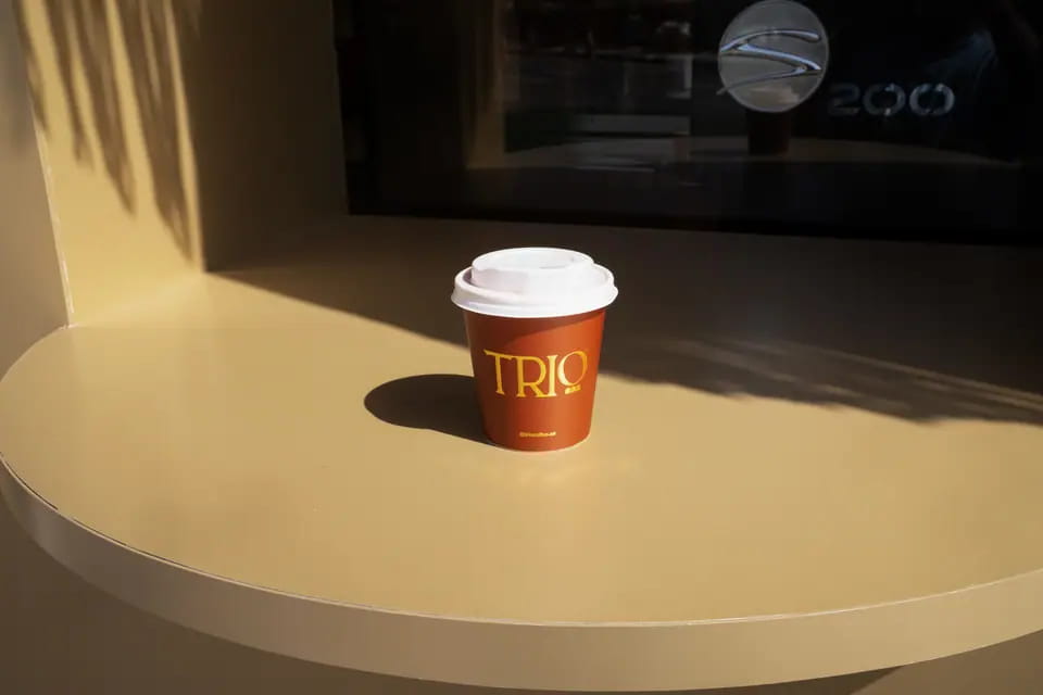 A terracotta TRIO takeaway cup on a curved sand-coloured counter in hard sunlight