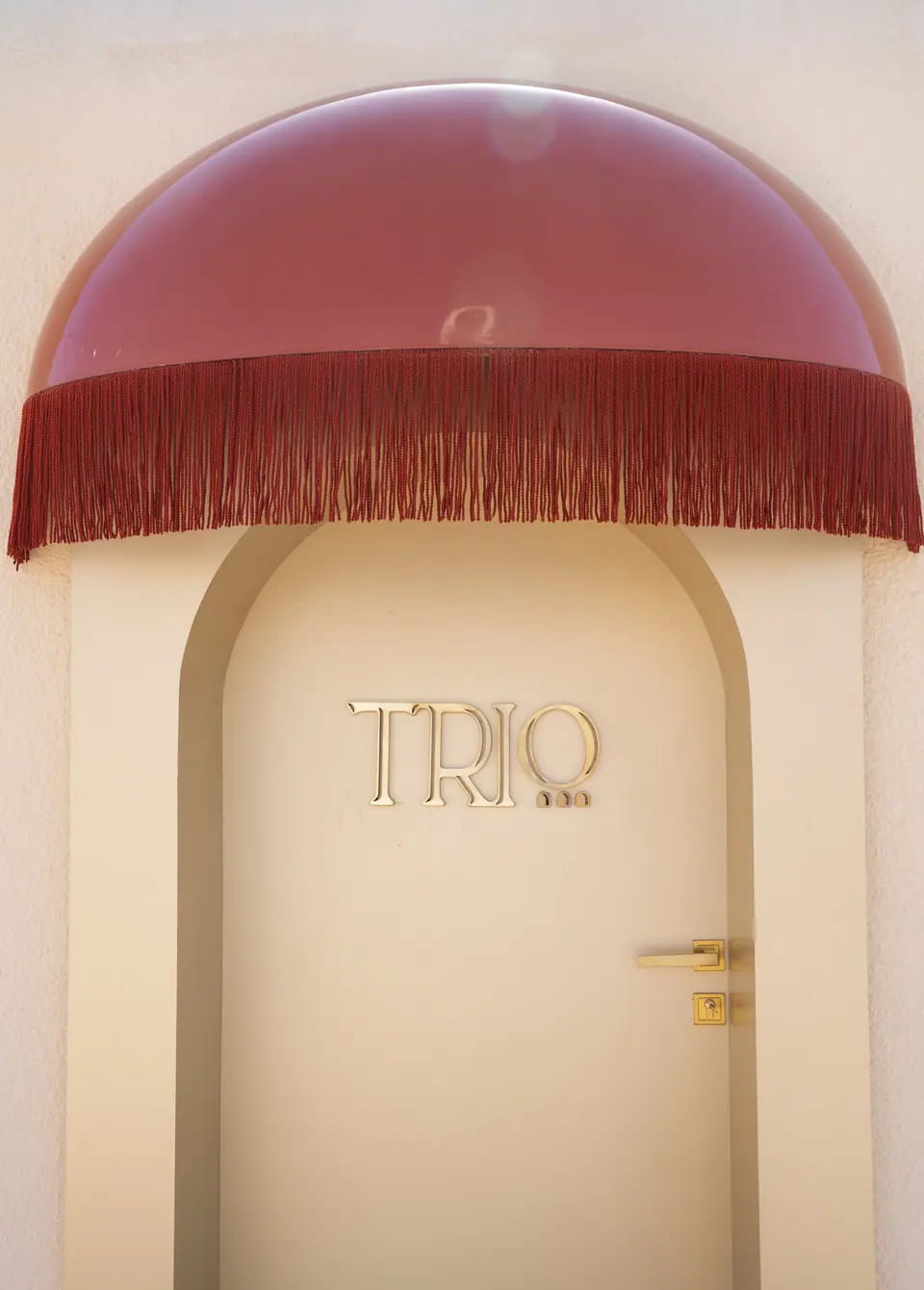 An arched cream door with a small brass TRIO wordmark under a glossy terracotta dome awning trimmed with long fringe
