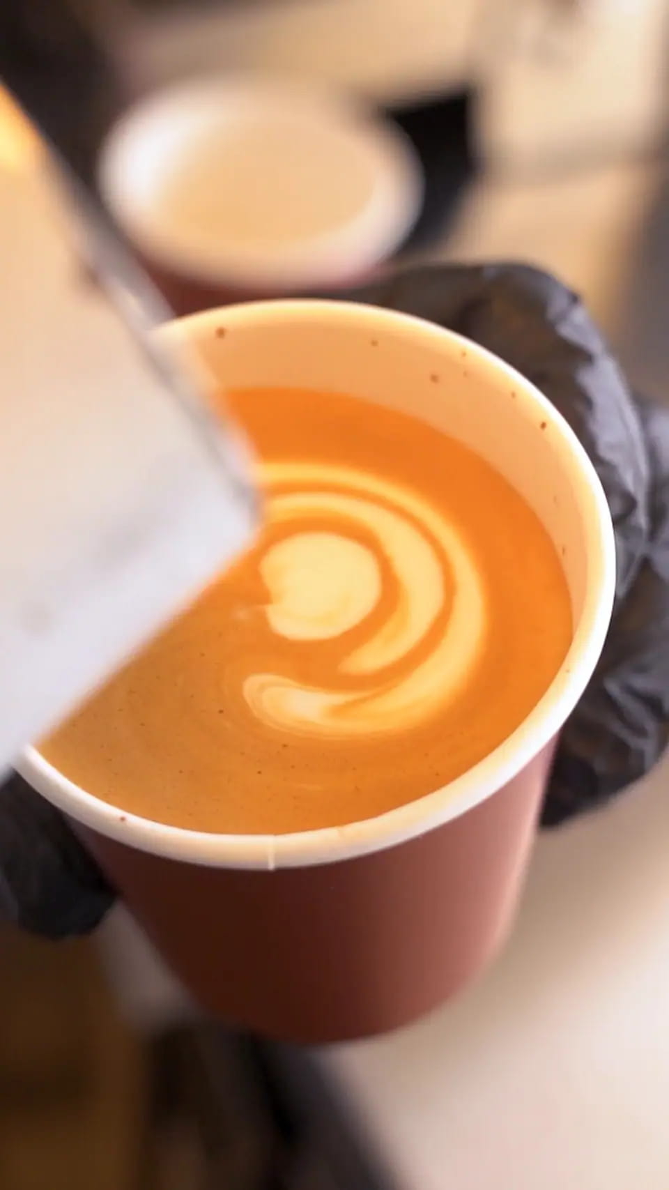 Milk poured into an espresso in a paper takeaway cup held in a gloved hand, forming layered latte art