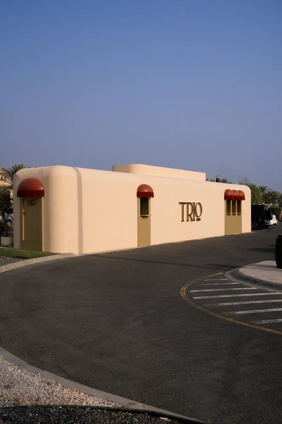 TRIO's rounded cream stucco building at Last Exit Al Khawaneej, a gold wordmark on its side and small red fringed awnings, under a clear blue sky