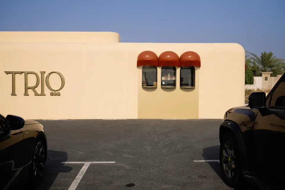 A long cream wall with a large brass TRIO wordmark and a trio of red-awninged service windows across the car park