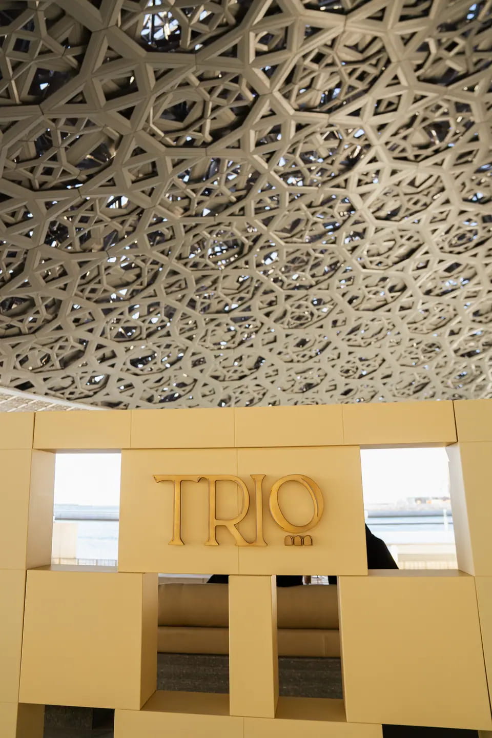 TRIO at Louvre Abu Dhabi: a cream sign gate of stacked blocks with square window cut-outs and gold TRIO lettering, under the museum's star-patterned dome