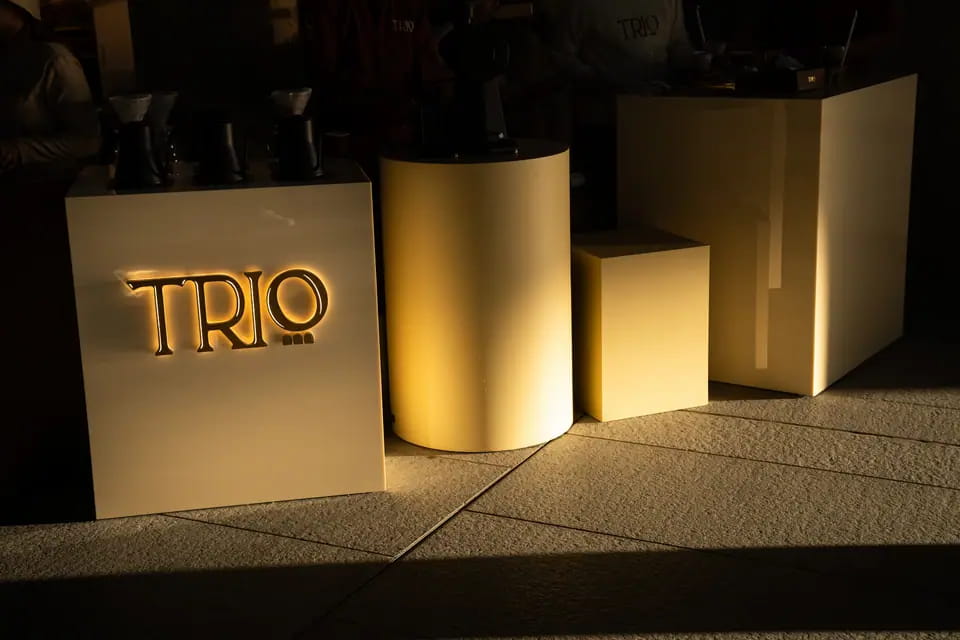 Low sun rakes across cream plinths and a brass cylinder at the Louvre bar, a backlit TRIO wordmark glowing on the front block
