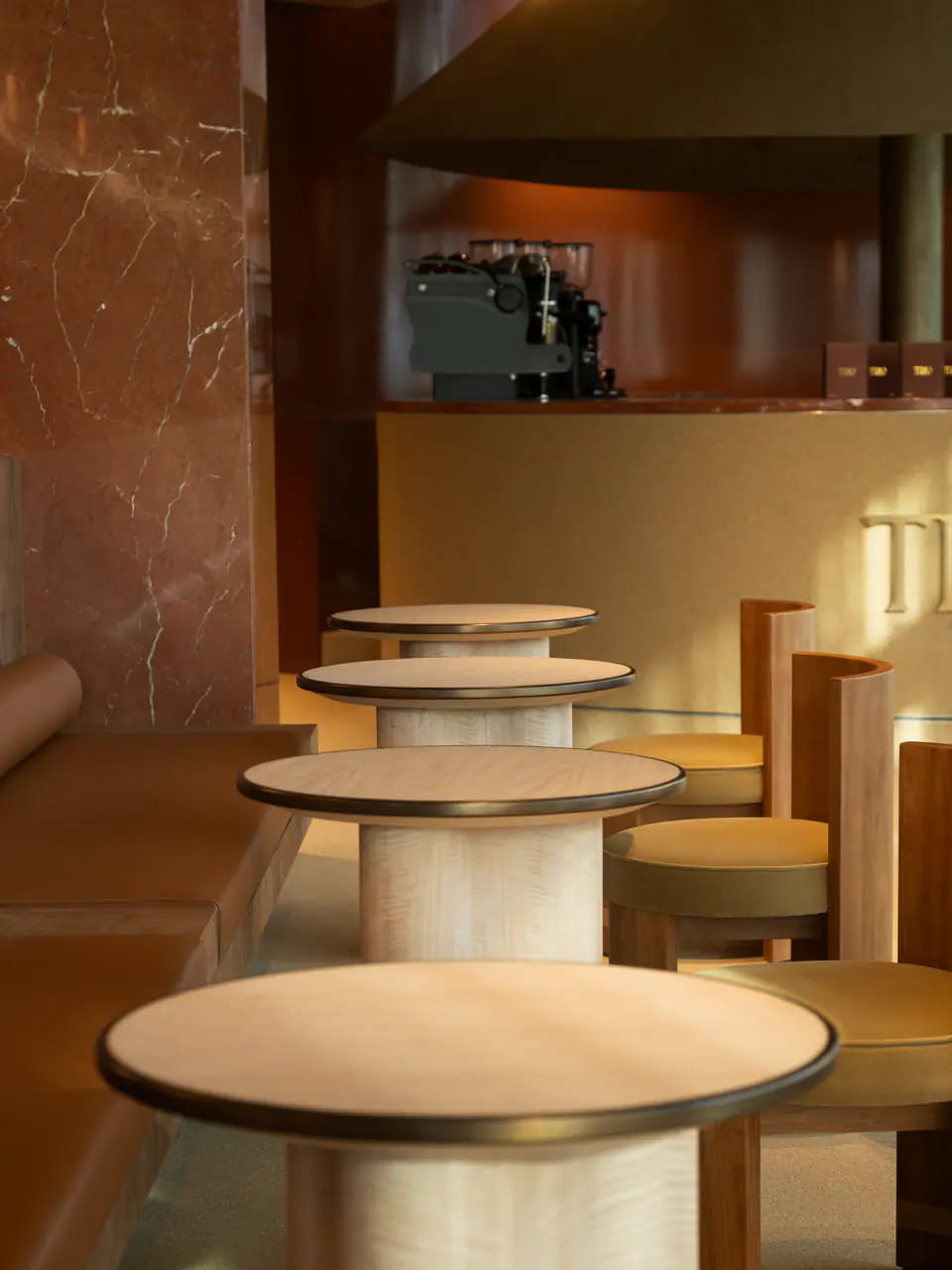 Round maple tables with brass rims in a row beside a leather banquette and red marble column, the espresso machine on the bar behind
