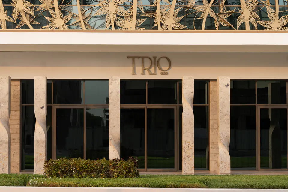 TRIO's travertine shopfront at Mirdif 35: sculpted stone pillars between tall bronze-framed windows, a brass TRIO wordmark on the white fascia and a gilded palm screen above