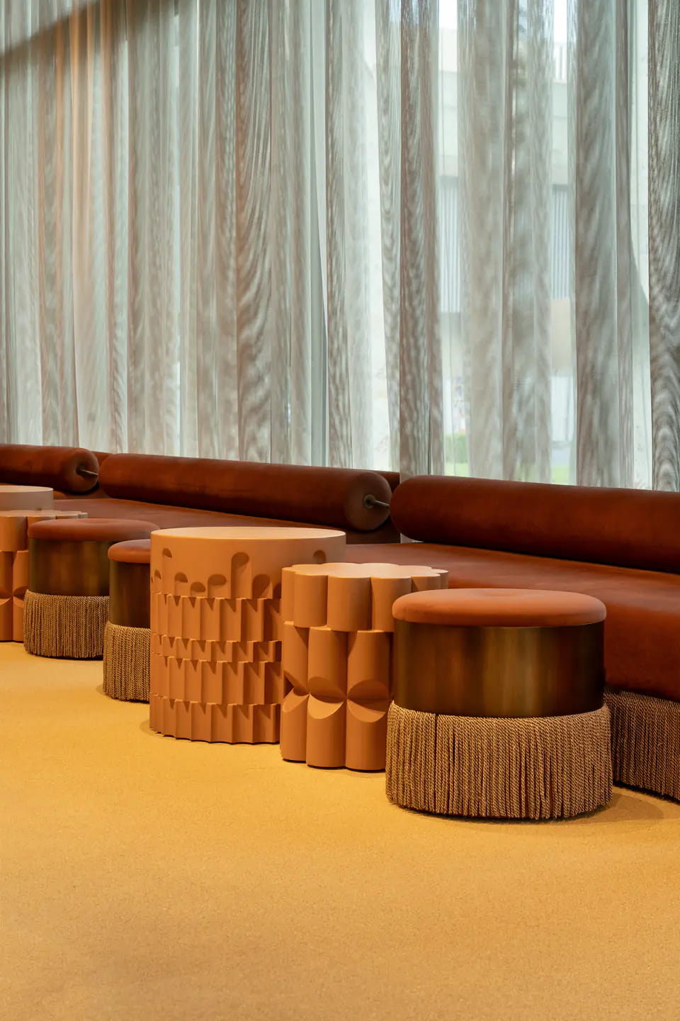 Terracotta pouffes with scalloped sides and fringed skirts along a rust leather bench, sheer curtains behind