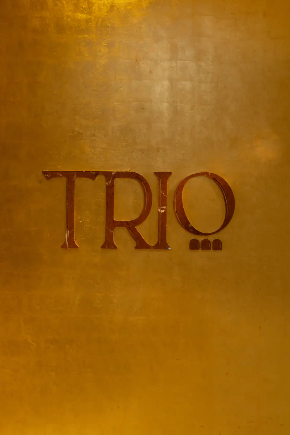 The TRIO wordmark in deep red on a wall of burnished gold leaf