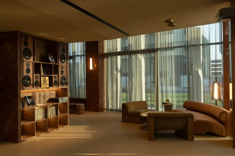 A listening corner with a walnut shelf of speakers, a turntable amplifier and records, beside low bouclé sofas and a stone coffee table in late sun through sheer curtains