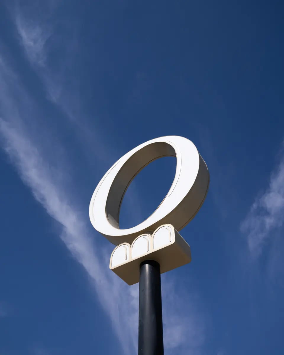 TRIO's cream O sign on a black pole, seen from below against blue sky and long streaks of cloud