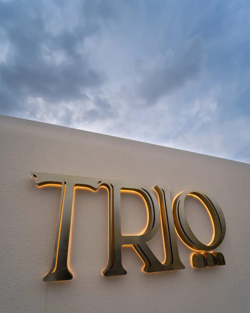 The brass TRIO wordmark backlit in gold on the wall of TRIO Umbrella Beach, Fujairah, against a grey sky