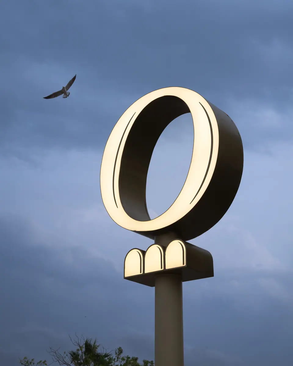 TRIO's illuminated brass O-and-arches pylon against a blue storm sky, a gull passing