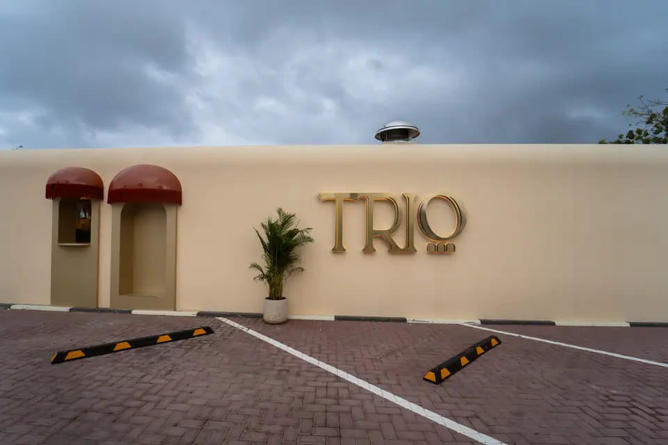 The long cream wall with two fringed awnings, a potted palm and the brass TRIO wordmark, looking across the brick-paved car park