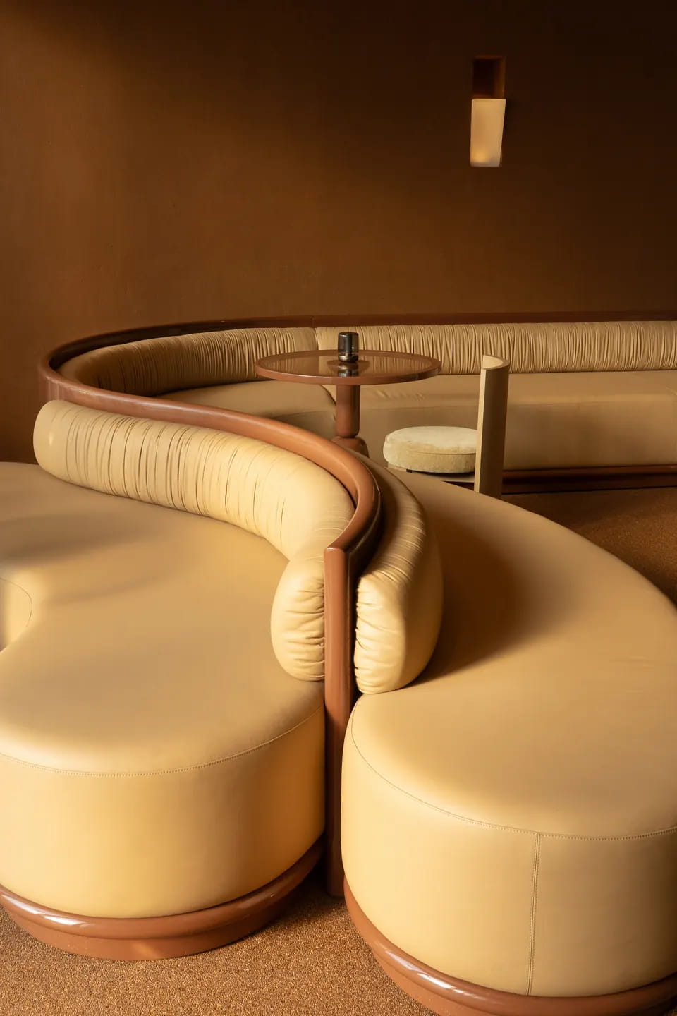 A cream leather banquette curves in an S around a round table, edged in glossy chestnut wood, a slim sconce glowing on the brown plaster wall