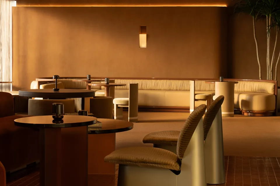 A long cream banquette runs the length of a brown plaster wall under a single sconce, with dark round tables and ochre shell chairs in front