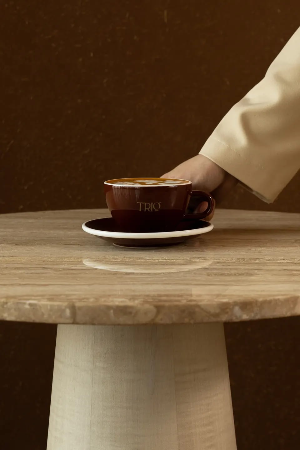 A hand sets down a brown TRIO cup of latte on a travertine table against a warm brown wall