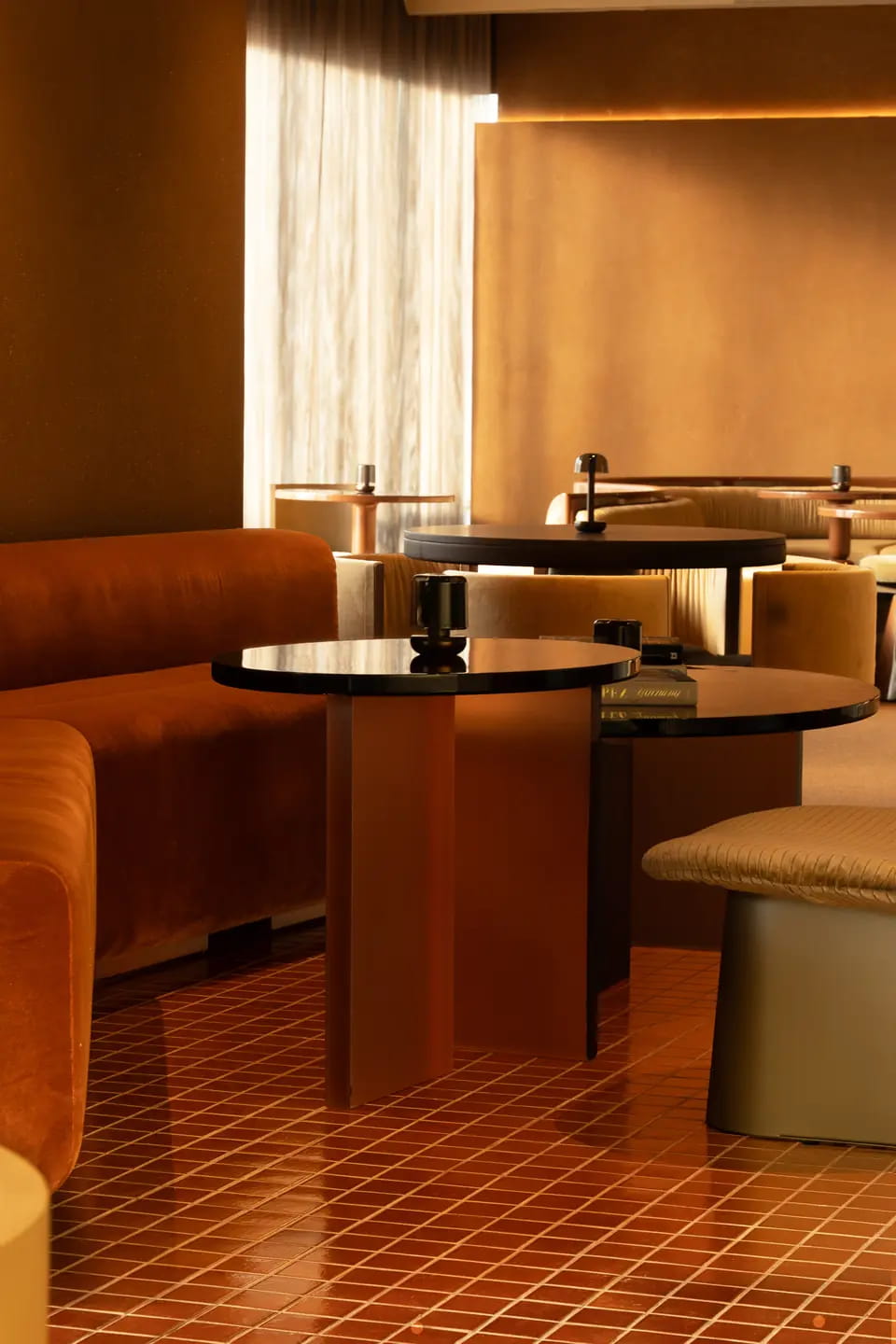 Rust velvet booths and dark glass-topped tables on a glazed terracotta tile floor, lit by a strip of daylight through the curtains