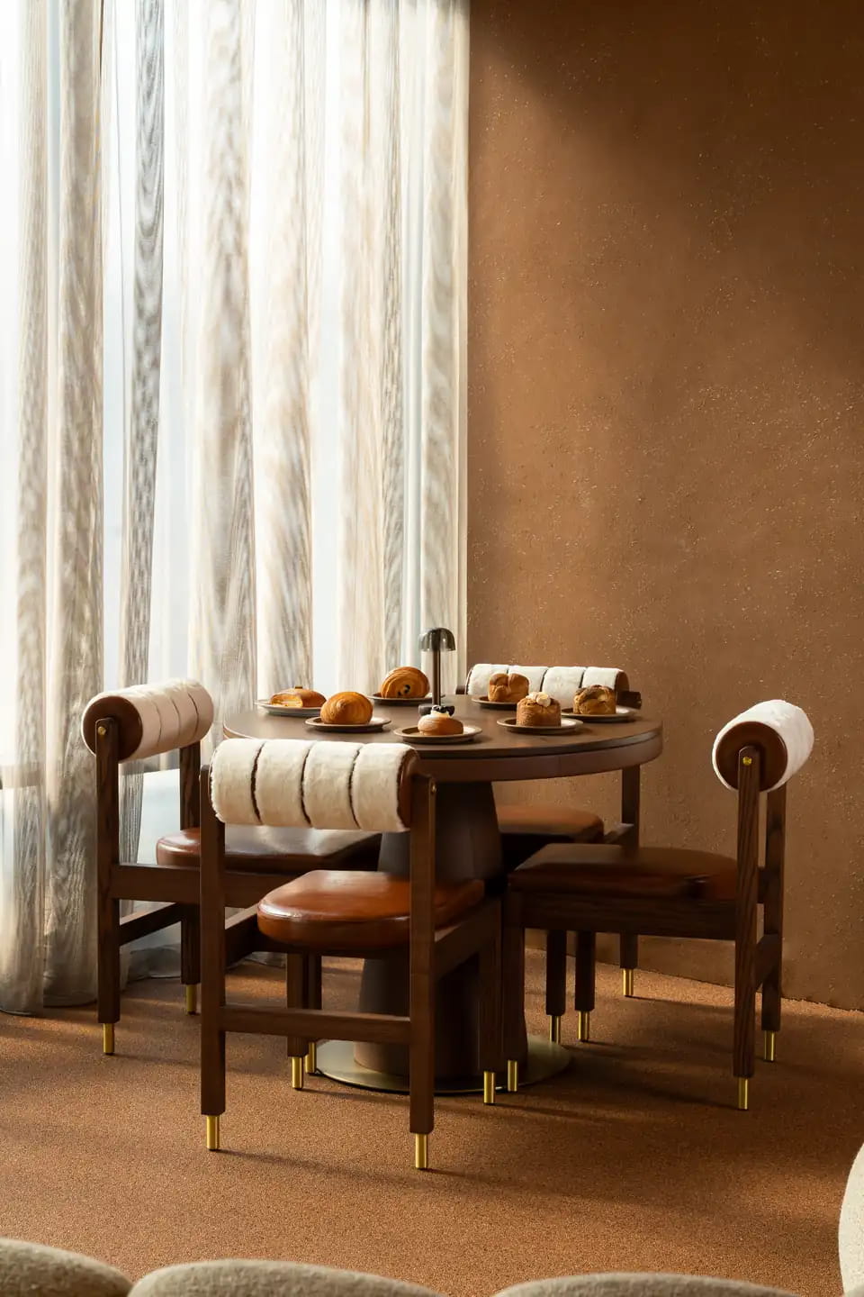 A round walnut table laid with pastries and coffee by sheer curtains, its chairs wrapped in cream fleece with brass-tipped legs