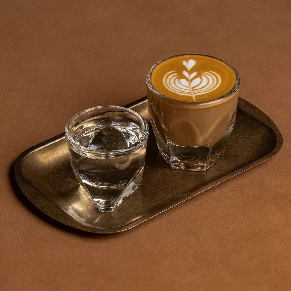 Cortado with latte art beside a glass of water on a brass tray