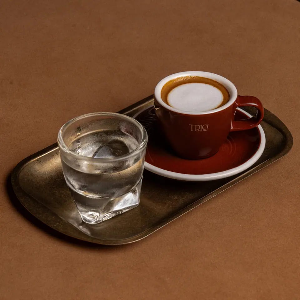 Macchiato in a burgundy TRIO cup with water on a brass tray